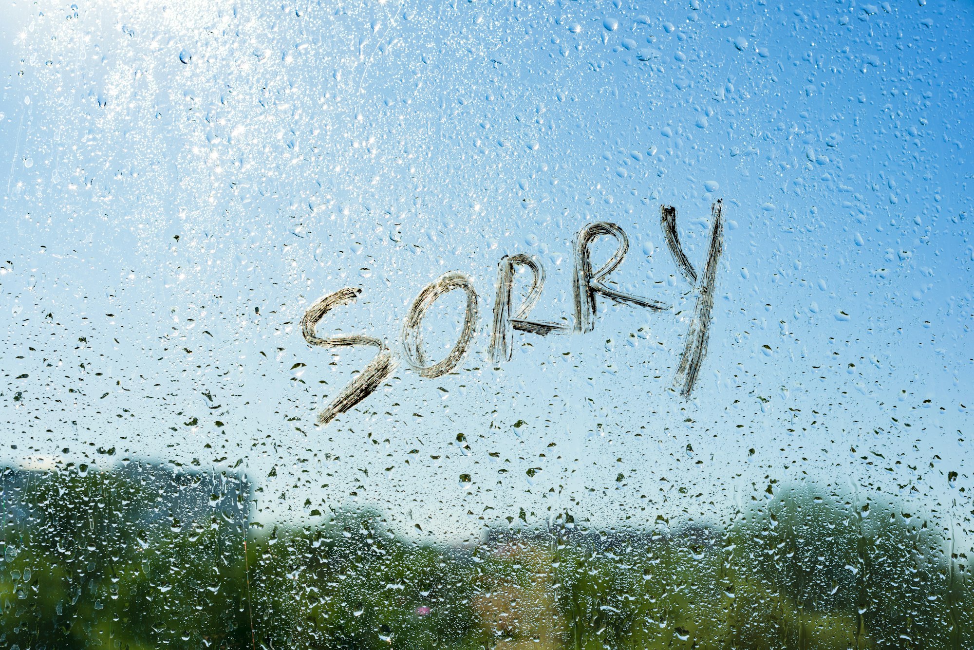 Word sorry on the window with raindrops. Background drop of water on the glass, blue sky, sun, city.